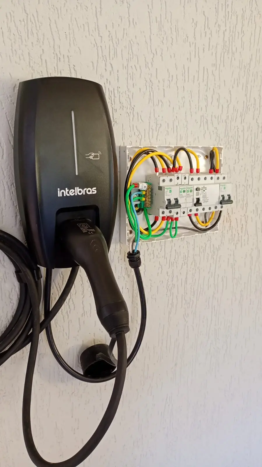 Carregador de veículo elétrico na parede, ao lado de caixa com disjuntores.