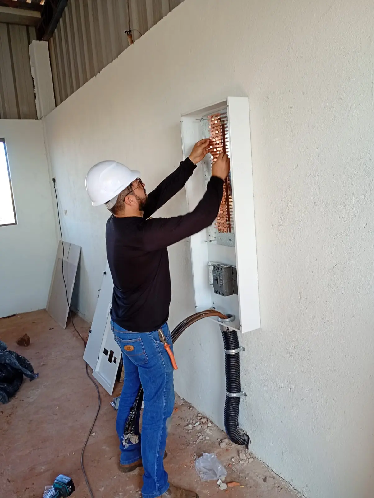 Eletricista com capacete executando instalação em quadro elétrico na parede.